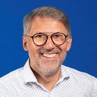 Portrait d'un homme souriant avec lunettes sur fond bleu.