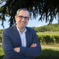 Homme souriant avec lunettes et veste devant un vignoble.