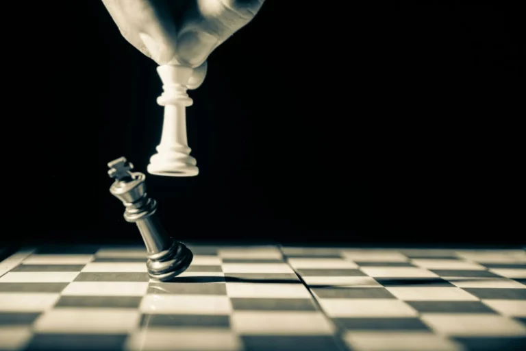 Une main déplaçant une pièce d'échecs blanche pour abattre une pièce noire sur un échiquier, sous un éclairage dramatique.