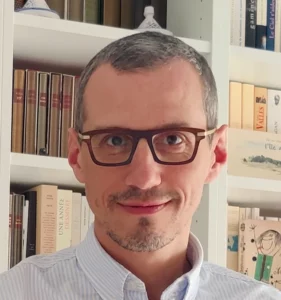 Portrait souriant d’un homme portant des lunettes, devant une bibliothèque remplie de livres variés.