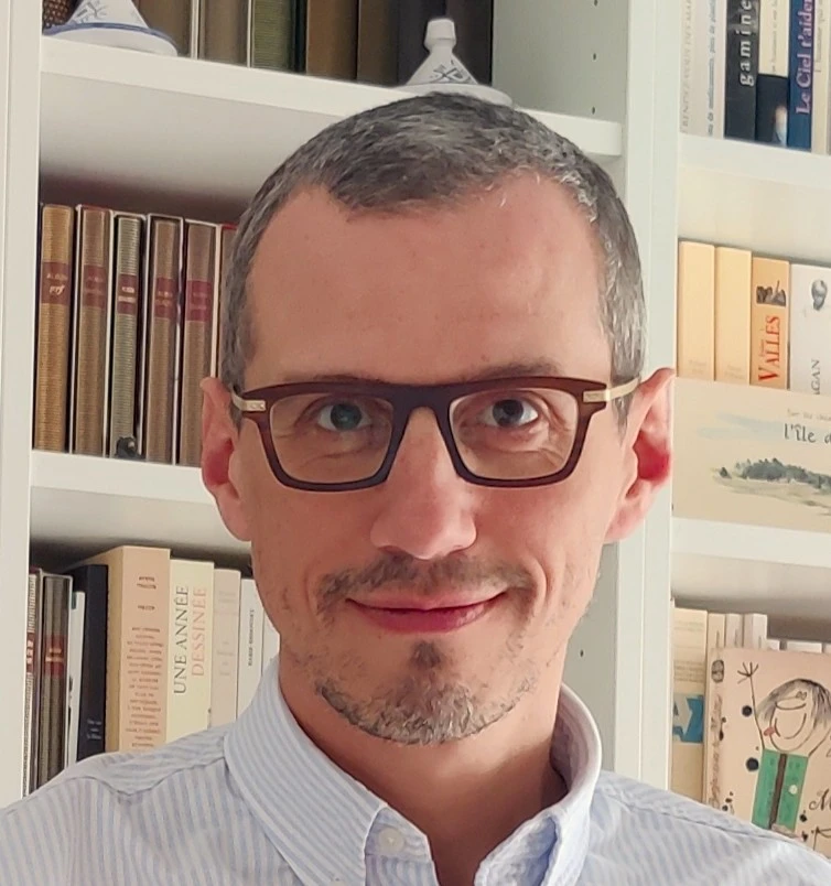 Portrait souriant d’un homme portant des lunettes, devant une bibliothèque remplie de livres variés.